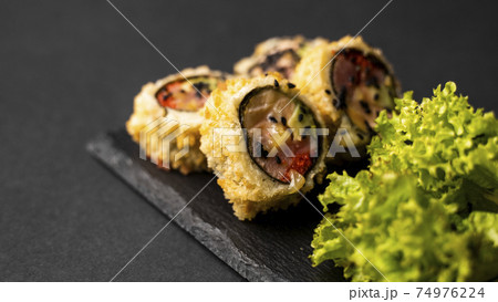 Custom sushi roll in tempura with nori, fresh salmon, tuna, avocado, masago caviar, drizzled with pineapple sauce with salad pouring as decoration on a black plate on a black table and background. 74976224