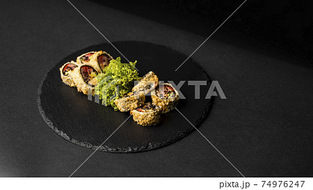 Custom sushi roll in tempura with nori, fresh salmon, tuna, avocado, masago caviar, drizzled with pineapple sauce with salad pouring as decoration on a black plate on a black table and background. 74976247