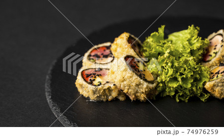 Custom sushi roll in tempura with nori, fresh salmon, tuna, avocado, masago caviar, drizzled with pineapple sauce with salad pouring as decoration on a black plate on a black table and background. 74976259