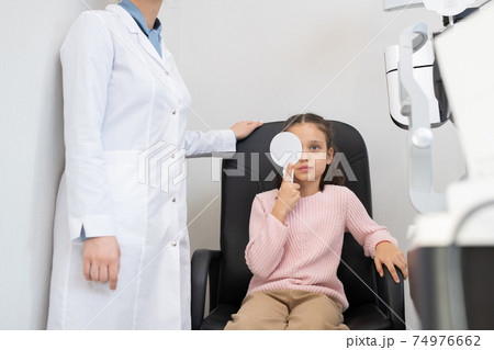 Cute little girl in casualwear doing eye test in modern ophthalmological clinics 74976662