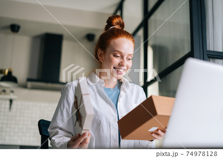 Low-angle shot of cheerful redhead young woman opening gift box with present during video call 74978128