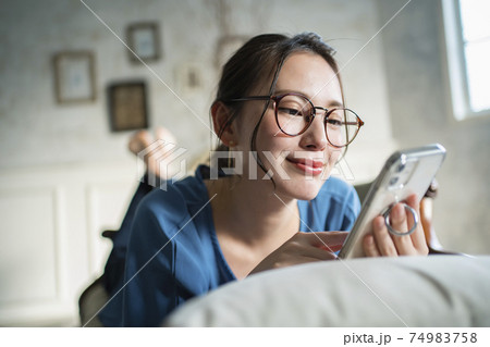 ソファに寝そべってスマホを操作する若い女性 74983758