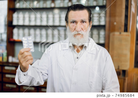 Take care of yourself, take medicine. Portrait of confident likable senior pharmacist in his ancient pharmacy, holding blister pack with white pills 74985684