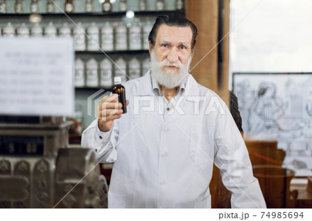 Portrait of a cheerful handsome elderly man pharmacist standing at the counter at old vintage drugstore and holding bottle with drops or syrup Portrait of a cheerful handsome elderly man pharmacist standing at the counter at old vintage drugstore and holding bottle with drops or syrup 74985694