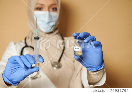 Muslim doctor with covered head holding syringe and vaccine. The concept of immunization of the population and the preventive fight against infections. Close-up Muslim doctor with covered head holding syringe and vaccine. The concept of immunization of the population and the preventive fight against infections. Close-up 74985794