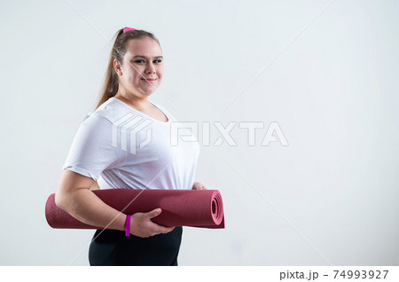 Young fat caucasian woman holding a sport mat. Charming plus size model in sportswear stands on a white background 74993927