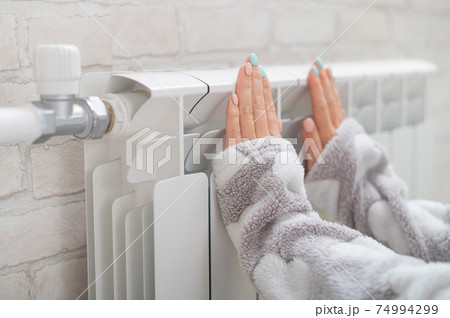 A woman wearing in pajamas warms cold hands on the heating radiator 74994299