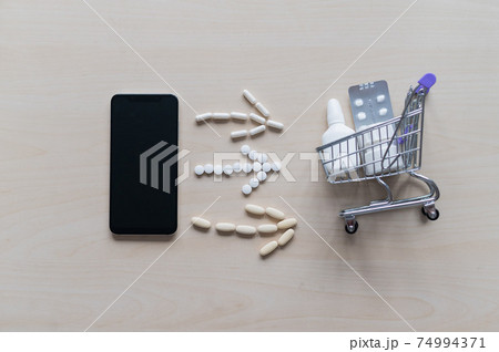 Smartphone app for the purchase of medicines. Top view on flat lay. A mobile phone with a black screen, tablets arrows and a mini shopping cart full of medications. Online pharmacy with home delivery. Smartphone app for the purchase of medicines. Top view on flat lay. A mobile phone with a black screen, tablets arrows and a mini shopping cart full of medications. Online pharmacy with home delivery. 74994371