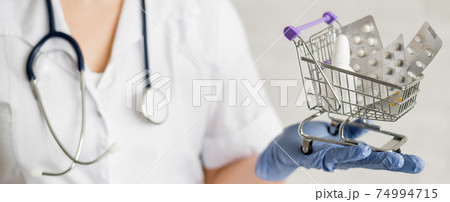 A faceless doctor with a medical coat and a stethoscope holds a small shopping cart full of medicines. Pharmacy concept. A faceless doctor with a medical coat and a stethoscope holds a small shopping cart full of medicines. Pharmacy concept. 74994715