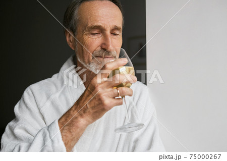 Close up of smiling mature man wearing white bathrobe holding wineglass looking away	 75000267
