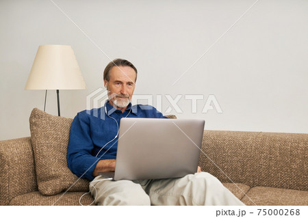 Senior man wearing earphones sitting on a couch with laptop	 75000268