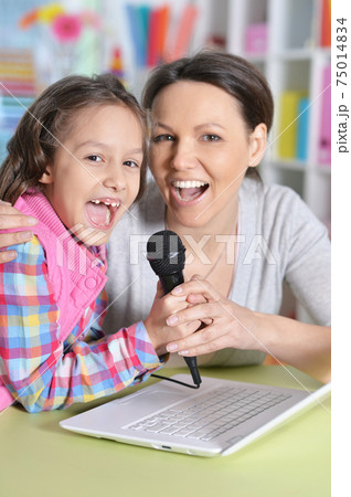 Cute little girl sitting at table and singing karaoke with mother 75014834