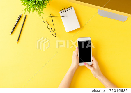 Overhead shot of female hands using smartphone with blank screen on yellow background with business accessories. Office desktop. Flat lay Overhead shot of female hands using smartphone with blank screen on yellow background with business accessories. Office desktop. Flat lay 75016682