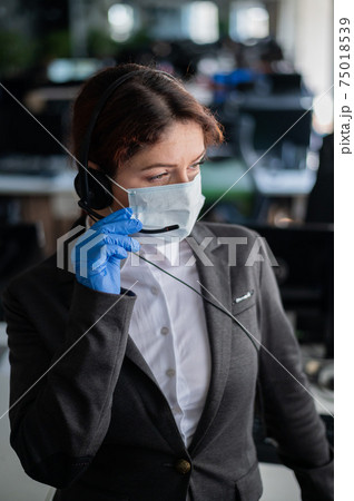 Female call center operator talking to a customer. Woman in mask and gloves at the workplace in the office. Social distance and safety measures to prevent infection. Female call center operator talking to a customer. Woman in mask and gloves at the workplace in the office. Social distance and safety measures to prevent infection. 75018539
