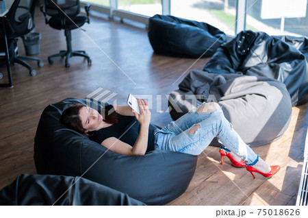 Caucasian business woman in casual wear lying and uses a smartphone in the lounge zone. Smiling girl looks at the mobile while sits in a beanbag in a modern office. A break at work. 75018626