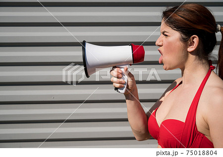 Red-haired Caucasian woman in a red bikini shouting through a megaphone against a background of a gray corrugated wall. A girl in a swimsuit is standing in profile with a loudspeaker. Summer vacation. 75018804