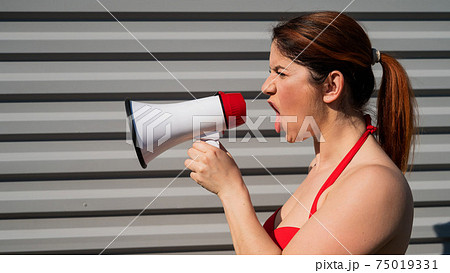 Red-haired Caucasian woman in a red bikini shouting through a megaphone against a background of a gray corrugated wall. A girl in a swimsuit is standing in profile with a loudspeaker. Summer vacation. Red-haired Caucasian woman in a red bikini shouting through a megaphone against a background of a gray corrugated wall. A girl in a swimsuit is standing in profile with a loudspeaker. Summer vacation. 75019331