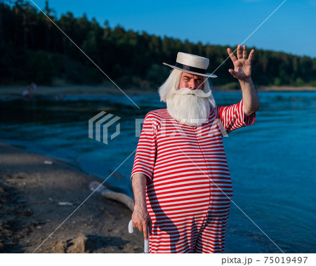 An elderly gray-haired man in a classic bathing suit and hat stands on the beach with an umbrella and waves his hand in greeting. Retired with a beard on vacation. An elderly gray-haired man in a classic bathing suit and hat stands on the beach with an umbrella and waves his hand in greeting. Retired with a beard on vacation. 75019497