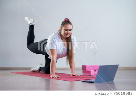 Young woman doing sports exercises at home. The girl is engaged in fitness at home on the mat and watches a training video on a laptop 75019985