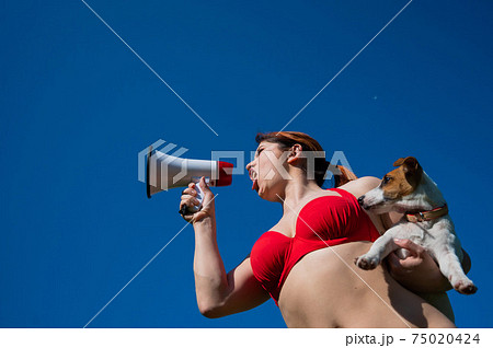 Caucasian woman in red bikini works as a lifeguard on the beach and shouts through a megaphone against a blue sky. Girl holds puppy jack russell terrier and loudspeaker. Summer vacation. Caucasian woman in red bikini works as a lifeguard on the beach and shouts through a megaphone against a blue sky. Girl holds puppy jack russell terrier and loudspeaker. Summer vacation. 75020424