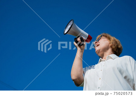 An emotional elderly woman pushes demands into a megaphone. An angry retired woman is fighting for the rights of older people. The female leader of the rally voiced claims to the loudspeaker. 75020573