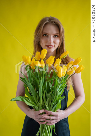 Caucasian woman with an armful of yellow tulips on a yellow background. International Women's Day. Bouquet of spring flowers 75020574