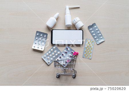 Online pharmacy concept with home delivery. Buying medicine online by phone. Mini trolley with different tablets, sprays, capsules and a smartphone with a white blank screen. Arrow made of pills. Online pharmacy concept with home delivery. Buying medicine online by phone. Mini trolley with different tablets, sprays, capsules and a smartphone with a white blank screen. Arrow made of pills. 75020620