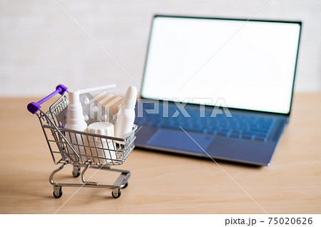 Laptop with blank white screen and miniature shopping cart full of medicines. Online pharmacy concept. Website for buying tablets. 75020626