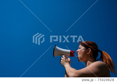 Red-haired Caucasian woman yelling into a megaphone against a blue sky background. The girl makes an announcement through the loudspeaker. Red-haired Caucasian woman yelling into a megaphone against a blue sky background. The girl makes an announcement through the loudspeaker. 75020628