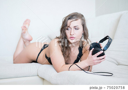 Beautiful woman in black lace underwear lies on her stomach and listens to music. The girl in the bedroom relaxes the bed holds headphones in her hands. 75020758