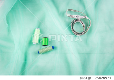 Tailor's inch, centimeter tape and green threads on a background of green tulle fabric. Tailor's equipment. Sewing tools. Tailor's inch, centimeter tape and green threads on a background of green tulle fabric. Tailor's equipment. Sewing tools. 75020974