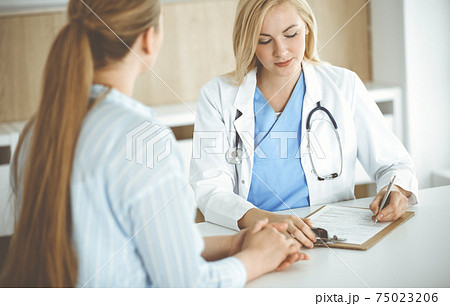 Woman-doctor and patient sitting and talking in hospital or clinic. Blonde therapist is cheerfully smiling. Medicine concept Woman-doctor and patient sitting and talking in hospital or clinic. Blonde therapist is cheerfully smiling. Medicine concept 75023206
