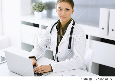 Woman-doctor typing on laptop computer while sitting at the desk in hospital office. Physician at work. Data in medicine and healthcare Woman-doctor typing on laptop computer while sitting at the desk in hospital office. Physician at work. Data in medicine and healthcare 75023815