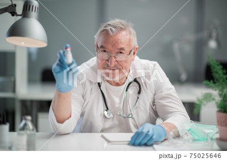 An elderly, experienced, reliable doctor demonstrates the coronavirus vaccine to the camera. The concept of vaccination of the population against covid-19 An elderly, experienced, reliable doctor demonstrates the coronavirus vaccine to the camera. The concept of vaccination of the population against covid-19 75025646