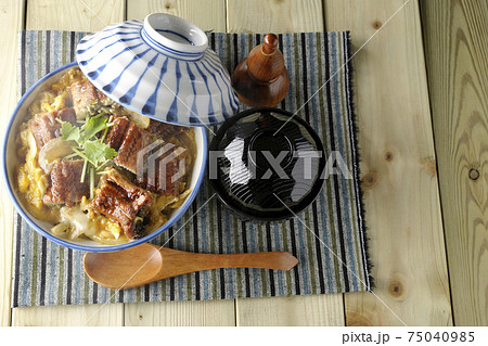 ランチョンマットの上の鰻蒲焼きを卵でとじた熱々うな玉丼とお吸い物のセットと木製スプーンを俯瞰で撮影 75040985
