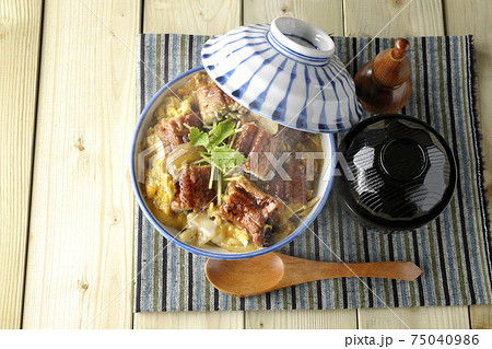 ランチョンマットの上の鰻蒲焼きを卵でとじた熱々うな玉丼とお吸い物のセットと木製スプーンを俯瞰で撮影 75040986