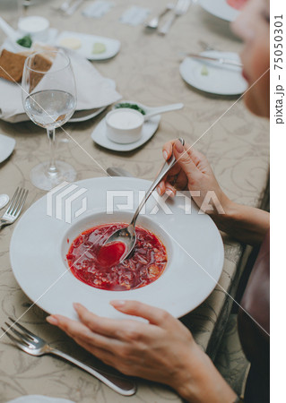 man eats red soup, sits at an elegantly served table in a restaurant, close-up. female hands with a spoon over red soup. borscht closeup man eats red soup, sits at an elegantly served table in a restaurant, close-up. female hands with a spoon over red soup. borscht closeup 75050301