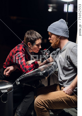 In the interior of a tattoo parlor, a young man with a beard is tattooed on his arm using the stick and poke method. Young artist girl in a checkered shirt. 75052553