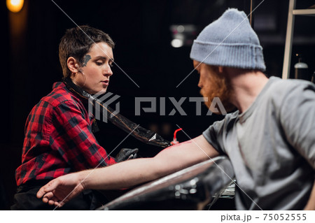 In a modern tattoo parlor, a girl with piercings and tattoos on her body makes a tattoo on the bicep of a young hipster's hand. 75052555