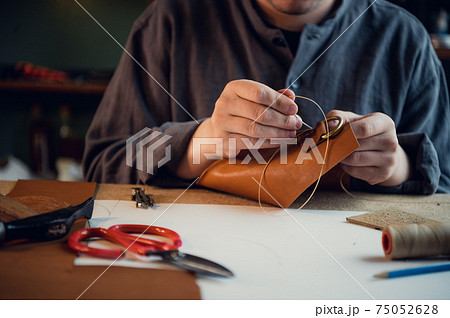 Sitting at a table in the workshop a young guy manually sews leather elements to each other 75052628