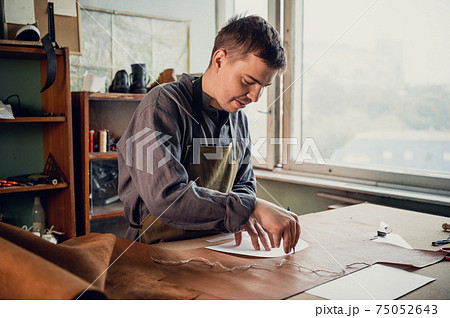 A young guy in a leather workshop transfers a pattern from paper to leather 75052643