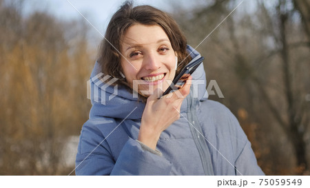 Young student in blue coat is using her smartphone to send a voice message to a new social network looking at the camera somewhere outdoors at the park. 75059549