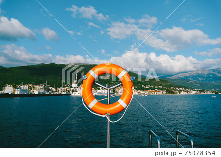Orange lifebuoy on a metal stand against the backdrop of mountains, water and sky. Orange lifebuoy on a metal stand against the backdrop of mountains, water and sky. 75078354