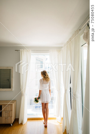 The barefoot bride in a white peignoir stands by the window with a wedding bouquet in her hands, back view The barefoot bride in a white peignoir stands by the window with a wedding bouquet in her hands, back view 75080455