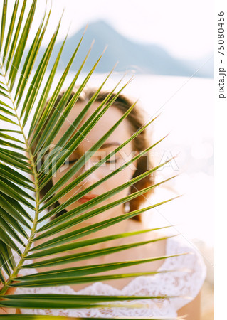 Portrait of a bride with closed eyes through a leaf of a palm tree, behind the sea and a mountain  75080456