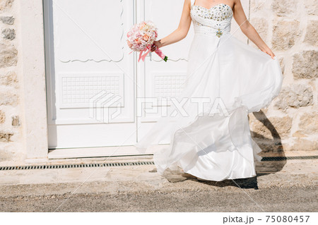 A bride in a stylish wedding dress and with a bridal bouquet stands at the beautiful white door of an old stone house A bride in a stylish wedding dress and with a bridal bouquet stands at the beautiful white door of an old stone house 75080457