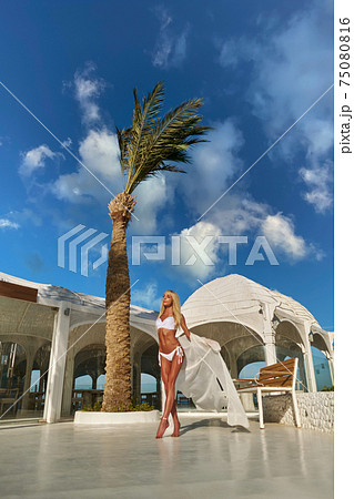 Attractive young woman wearing swimsuit and pareo standing over white exotic building 75080816