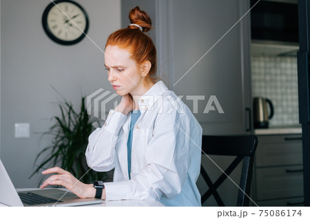 Tired redhead young business woman having neck pain during working at laptop compute Tired redhead young business woman having neck pain during working at laptop compute 75086174