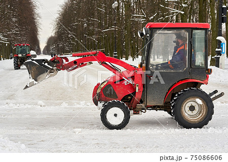 Red tractor with a large bucket removes snow in a city park Red tractor with a large bucket removes snow in a city park 75086606