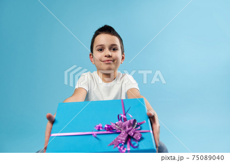 Cute little boy holds a blue gift with pink bow out to the camera while sitting on a blue background. Celebration Birthday concept Cute little boy holds a blue gift with pink bow out to the camera while sitting on a blue background. Celebration Birthday concept 75089040